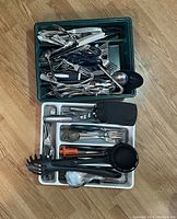 Top-down view of two plastic trays containing various used kitchen utensils including spoons, forks, peelers, tongs, can opener and spatula.