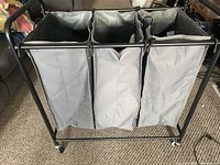 Front view of laundry cart showing three gray fabric bags attached to black metal frame with wheels on the floor beneath.