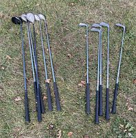 Photo showing nine golf clubs arranged on grass, including irons and woods, handles with black grips and metal shafts, heads with varied sizes.