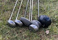 Two golf clubs lying on grass; a large wood club with black head marked '5' and a smaller silver iron club.