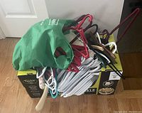 Cardboard box filled with assorted plastic, wooden, and metal hangers with a green reusable bag on top.
