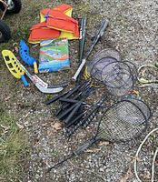 Photo showing all items together including water slide box, sports bats, rackets, paddles, hockey stick, life vest, and other accessories placed on gravel.