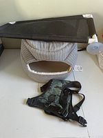 Grey and white patterned pet bed, camouflage dog harness, and black elevated pet cot all on a light floor surface.