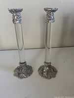 Pair of pewter candlestick holders with clear central columns and decorative grapevine patterned bases and tops displayed on white surface against a neutral background.