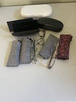Four pairs of reading glasses in various cases/pouches plus leopard print pouch displayed on flat surface.