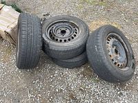 Four used tires mounted on steel rims arranged on gravel ground, showing some dirt and wear.