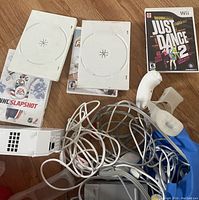 Photo showing Nintendo Wii console, two game cases NHL Slapshot and Just Dance 2, two white Wii Remote controllers with one silicone cover, and a bundle of cords on wood floor.