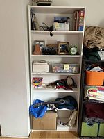 White IKEA Billy bookshelf with five shelves containing miscellaneous household items including framed photos, small boxes, toys, and a small blue guitar at the bottom.