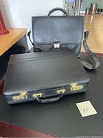 Two black leather briefcases on a black surface. One is a traditional briefcase with combination locks and a handle, the other a messenger-style briefcase with a flap and shoulder strap.