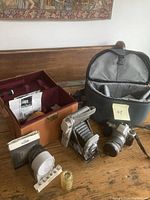 Pentax MZ-6 camera with attached 28-80mm zoom lens, flash unit, and carrying backpack along with Polaroid Land Camera 80A in original case with accessories on wooden surface