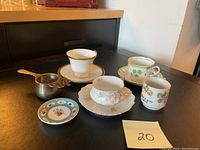 Four tea cups with three saucers, a small floral plate, silver tea strainer, and nine collector spoons arranged on a black surface.