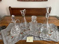 Four crystal decanters displayed on a table with an embroidered cloth. Two taller wine decanters on right and left, one with an octopus-shaped aerator. Two shorter whiskey/sherry decanters with pinwheel patterns center.
