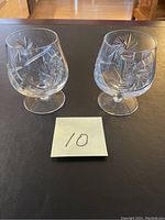 Two cut crystal brandy glasses with pinwheel design shown side by side on a dark surface with a note '10' in front. One glass appears intact, the other may have minor damage.