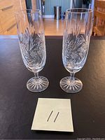 Pair of tall cut crystal champagne glasses with starburst pinwheel cut design shown standing side by side on a dark surface.