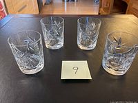 Four cut crystal whiskey glasses arranged in two rows on a dark tabletop with a note numbered 9 in the center, showing overall set condition and design.