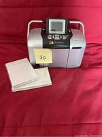 Photo of Epson PictureMate Deluxe 500 photo printer on red fabric surface, with stacks of photo paper beside it.