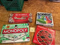 Four board games including Monopoly, Scrabble, Sorry, and Checkers visible in their boxes, displayed on a wooden floor.