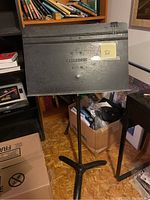 Photo shows a black metal music stand with 'MANHASSET' embossed on the front panel, a tripod base, adjustable pole, and a large binder clip on top right of the music resting area.