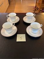 Four bone china tea cups and saucers arranged on a dark surface with a sticky note marked '5'. The cups and saucers have a consistent white and silver platinum design.