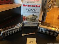 Photo showing all three KitchenAid pasta attachments (roller, fettuccini cutter, spaghetti cutter) and original box with product branding and imagery.
