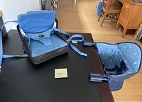 Both portable baby seats shown on a table; one blue seat hangs from table, the other blue patterned seat straps to a chair.