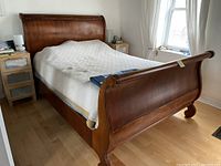 Full side view of queen size wooden sleigh bed showing curved headboard and footboard, mattress covered with white quilt, some scratches and wear visible.