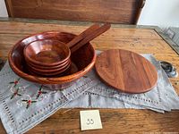 Photo showing large wooden salad bowl with six small bowls nested inside and two salad servers placed on top. A grey linen cloth with floral embroidery lies underneath on a wooden table.