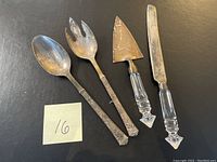 Four serving utensils arranged on black surface with visible tarnish and wear. Includes two ornate silver salad servers plus a pie server and a cake knife with glass handles.