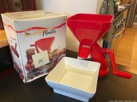 Red plastic tomato press with hand crank and white tray on kitchen counter next to original box.
