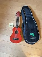 Full view of orange Emus ukulele laying on floor next to black carrying case with palm tree artwork