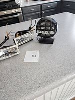Three 12 volt fog lights: two rectangular clear lens fog lights with wires and one round black PIAA fog light with mounting bracket, shown on kitchen counter.