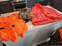 Camouflage jacket with orange lining, heavy orange safety vest with hood, lightweight orange safety vest, and camouflage cap laid out on a kitchen counter.