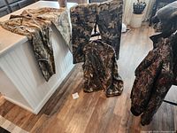 Camouflage hunting vest, jacket, and two pants laid out on wooden floor and kitchen counter.