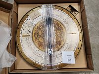 Photo of round decorative wall clock face with brass or gold-tone frame, wrapped in plastic and resting in a box.