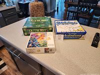 Photo showing the three items on a countertop: Outburst game in green box, Telestations game in blue and white box, and 1000 piece glitter fantasy puzzle with colorful artwork.