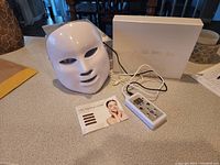 White LED facial mask placed on table with wired remote and instruction booklet visible, box in background.