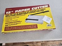 Box label showing product features and images of the 12 inch paper cutter including capacity for 10 sheets, lightweight steel base, fabric blade lock, rubber feet, and adjustable sliding ruler.