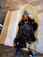 Full view of the doll lying in foam protective packaging inside a cardboard box with wrapping paper and a backdrop.