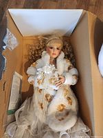 Porcelain doll with blonde hair dressed in white fur-trimmed coat with gold floral embroidery, white dress, and pearl accessories inside a cardboard box.