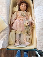 Full view of porcelain doll in original packaging showing entire figure dressed in pink with matching bow and shoes, along with protective foam.