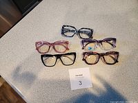 Photo showing five pairs of reading glasses arranged on a table including gray camouflage, pink, purple, black, and tortoiseshell frames.
