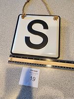 Square white metal hanging sign with black letter "S", shown hanging or laying flat next to measuring tape for size reference