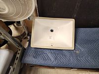 Side angle view of white ceramic under mount bathroom sink placed on padded surface showing overall rectangular shape and central drain hole.