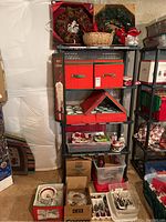 Photo of an assembled shelving unit stacked with two Christmas wreaths (pinecone berry and green with bells), wicker basket with red floral decor, red and green storage boxes, trays of flatware, plates, and glasses arranged on shelves and floor below.
