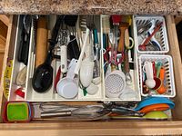 Top-down view of open drawer filled with assorted cooking utensils, measuring cups, spatulas, ladles, cutters and brushes all mixed in plastic white organizer trays and wooden drawer.
