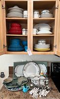 Photo showing open kitchen cabinet filled with stacked dinner plates, bowls in red, blue, white, yellow and Southwestern patterned dinnerware with flatware below on countertop.