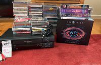 Full lot arranged on floor, showing black VHS player with remote, stacks of CDs, DVDs, and boxed VHS tapes including The X Files set and musicals.