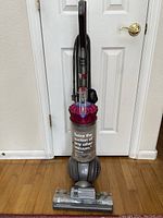 Full front view of Dyson upright vacuum cleaner showing clear dust bin and purple filter assembly on top, with cord attached to handle.