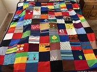 Overview photo of the large hand-knit patchwork blanket laid out on a bed, showing many colorful patches with different patterns and Colorado ski town names and logos.