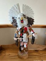 Front view of Kachina figure showing white feather headdress, yellow beak, large eyes, fur details, and painted traditional clothing.